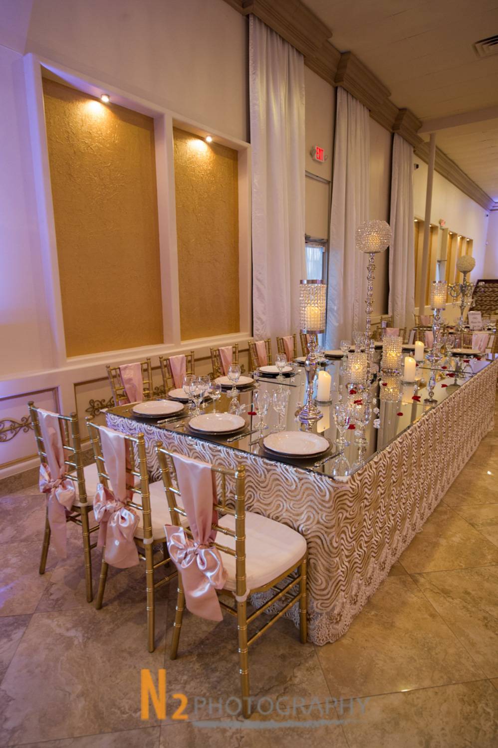 Elegant banquet table setup with mirrored tabletop, gold chairs adorned with pink satin bows, crystal candle holders, and fine china in a luxury event hall - Alegria Gardens - Houston, TX Elegant banquet table setup with mirrored tabletop, gold chairs adorned with pink satin bows, crystal candle holders, and fine china in a luxury event hall - Alegria Gardens - Houston, TX