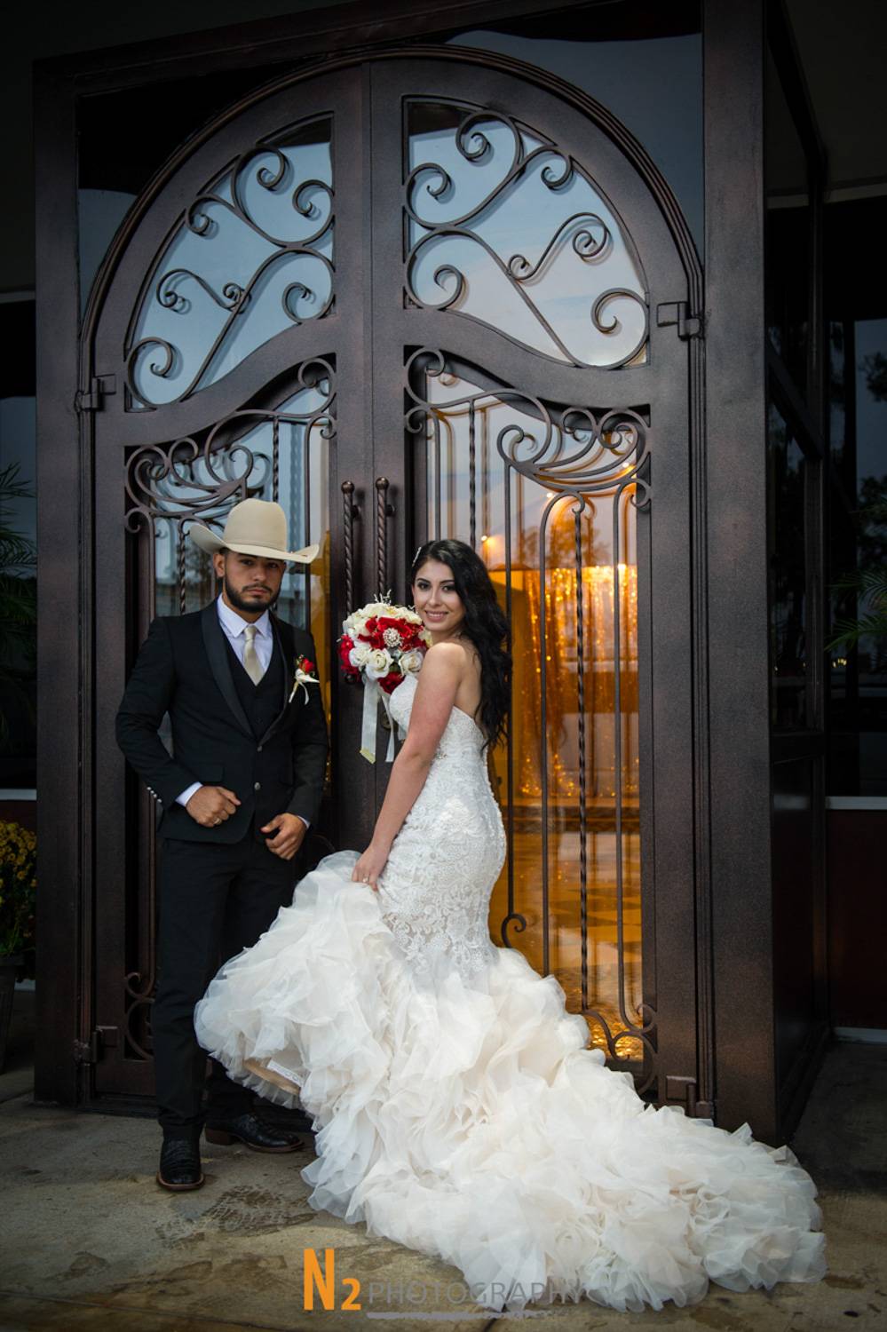 Bride and groom posing elegantly outside a venue with ornate wrought iron doors, showcasing timeless wedding style by Alegria Gardens event planners. Bride and groom posing elegantly outside a venue with ornate wrought iron doors, showcasing timeless wedding style by Alegria Gardens event planners.